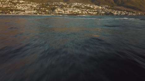 el dron vuela rápidamente sobre el mar ondulado en la playa de camps bay en ciudad del cabo, sudáfrica. muchas casas en la cima de una colina. vista de las rocas de la montaña en el mar.
