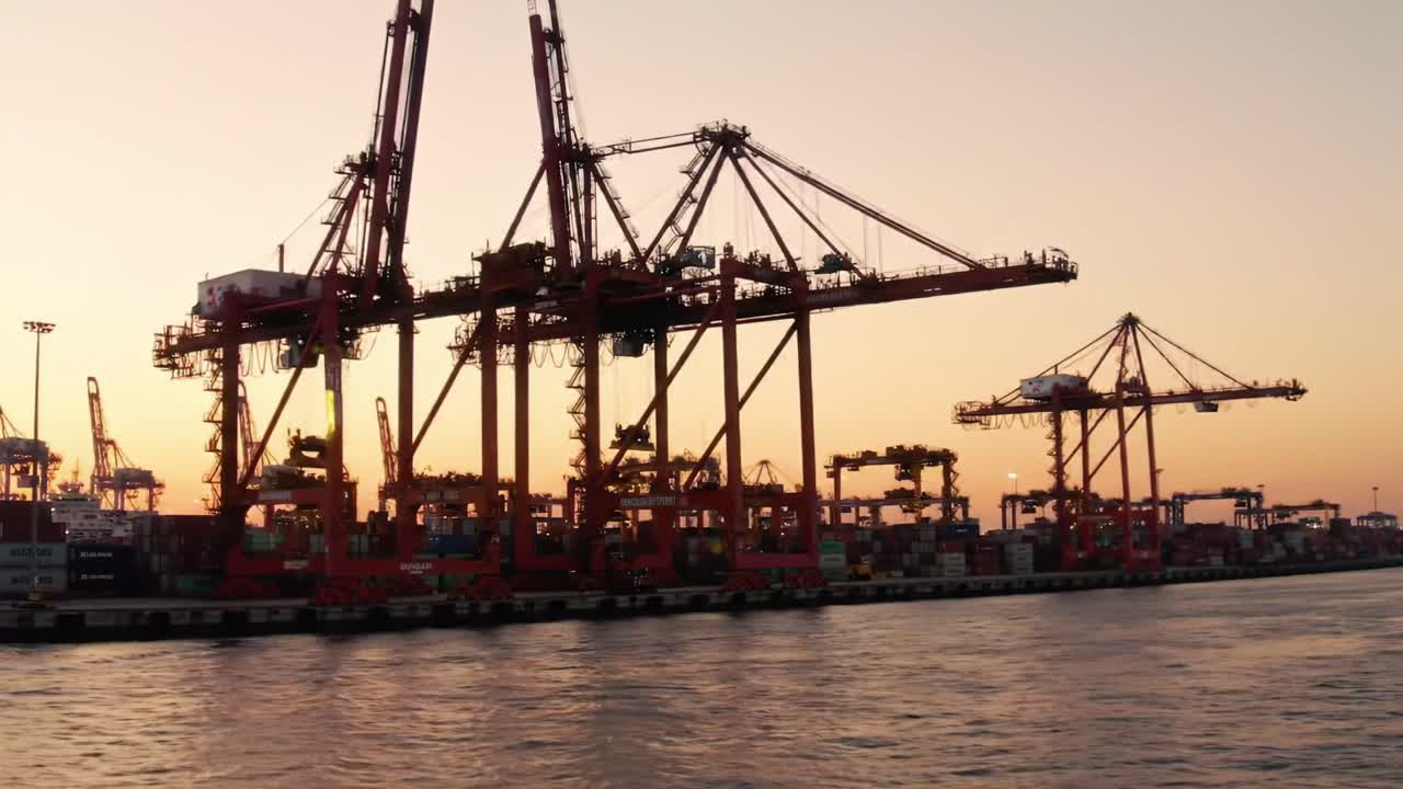 Sunset at the Port: Cargo Cranes Silhouetted Against a Golden Sky, Showcasing Maritime Industry in Action with Containers Lined Up Along the Water's Edge