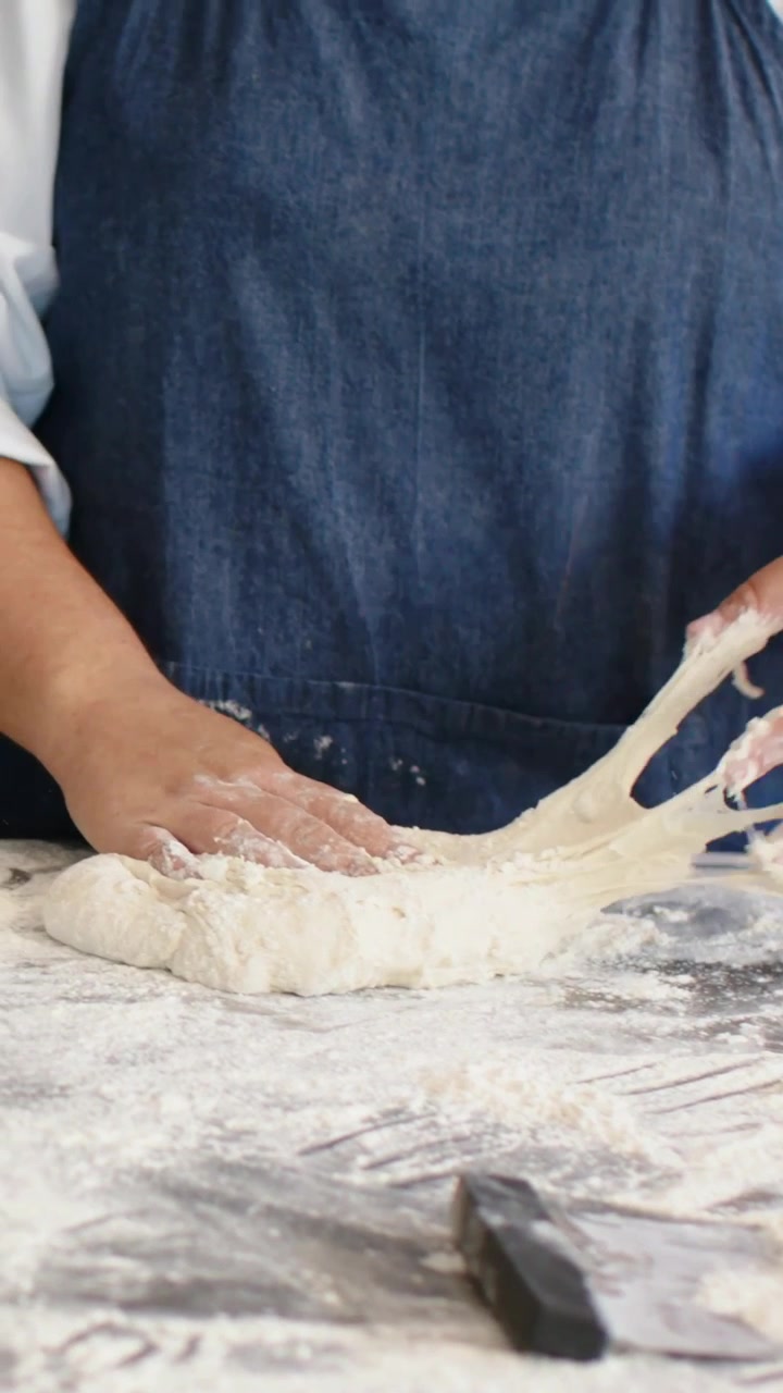 Vertical video: Camera focusing hands in bakery, chef in apron kneading dough for bread, scraper