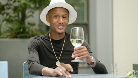 Happy-wealthy-man-wearing-fedora-hat-and-gold-jewelry-sits-at-a-table-drinking-wine-outside-near-green-plants
