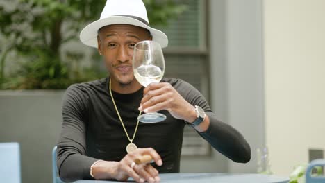 happy wealthy man wearing fedora hat and gold jewelry sits at a table drinking wine outside near green plants