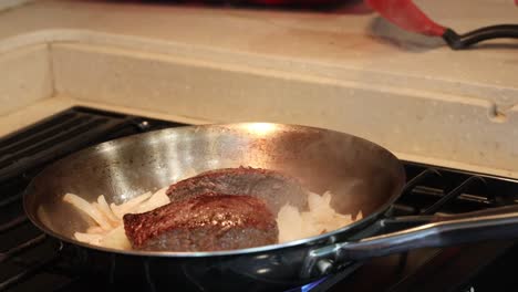 steaks and onions cooking in a stainless steel frying pan on a gas stove