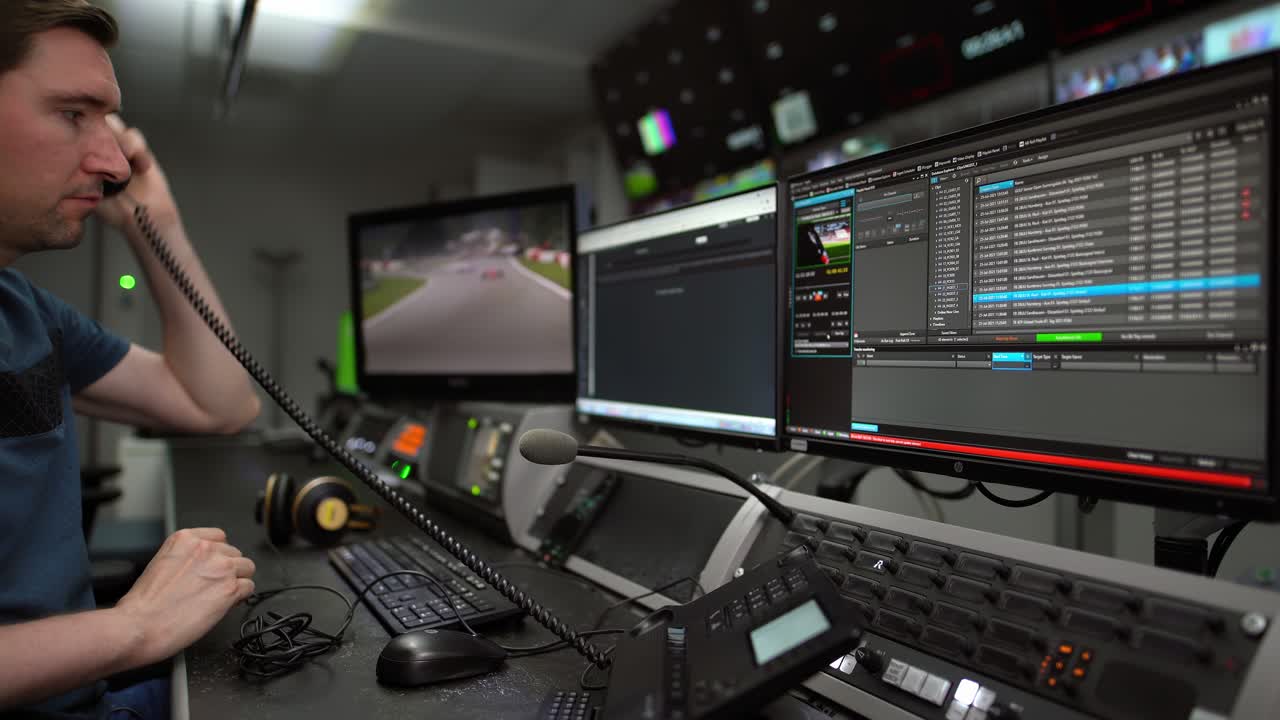 мужчина на рабочем месте в помещении телекомпании в наушниках, разговаривает по телефону и просматривает новости на компьютере