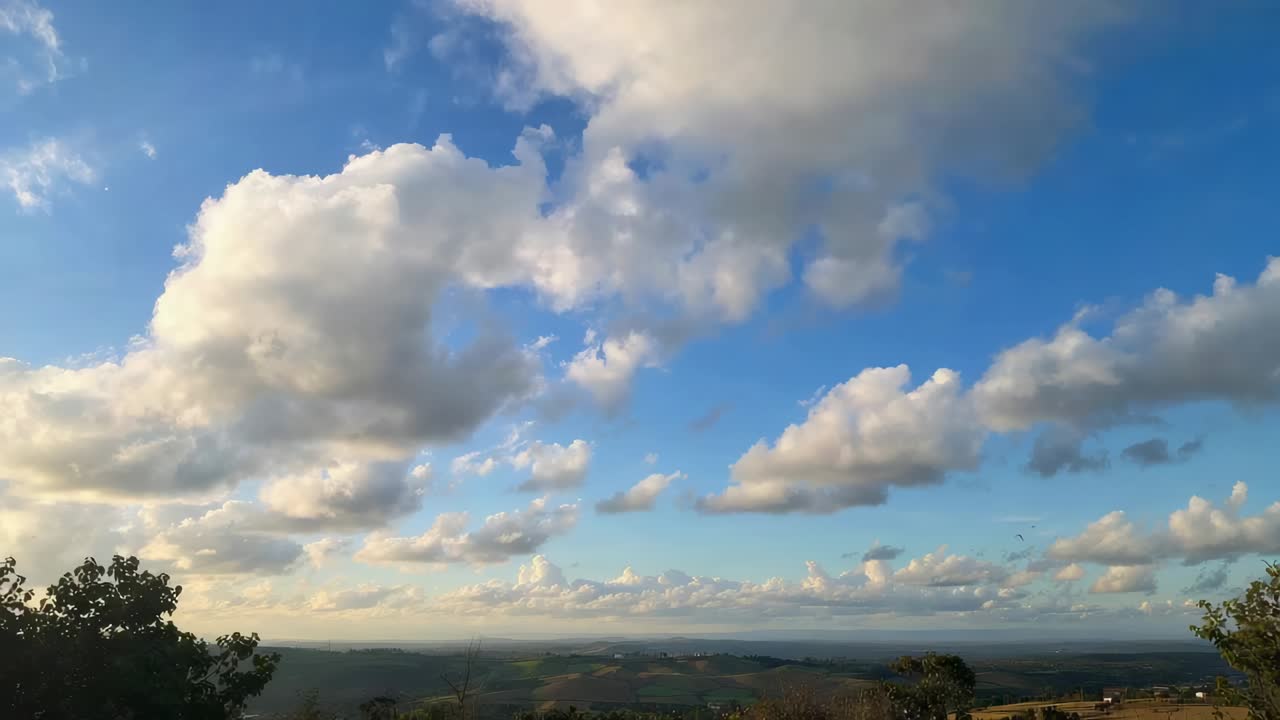 Nuvole cumulo alla deriva che si spostano nel cielo rurale, brezza che accarezza i bordi delle nuvole sopra colline e alberi.