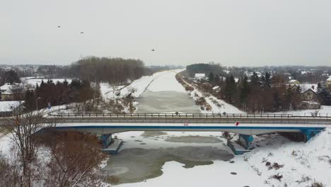 Drone-video-of-frozen-river-under-a-bridge-between-a-forest-covered-in-snow-in-warsaw