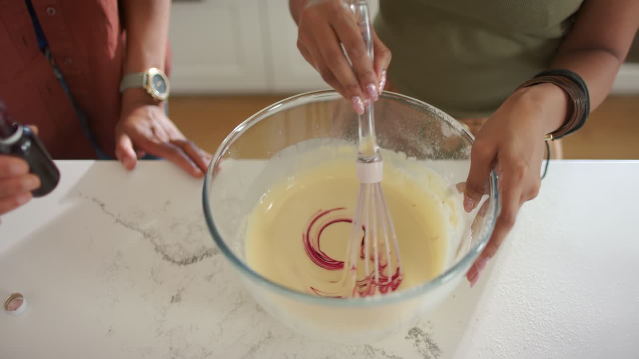 Indian and African American women tipping bottle and whisking in bowl on counter, coloring batter