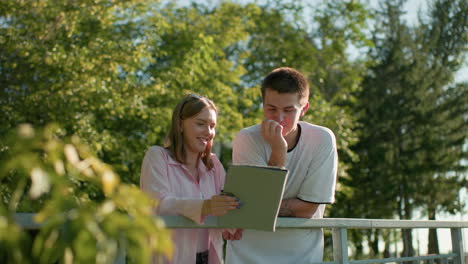 lady in pink top showing her brother something on her tablet while smiling and nodding as he laughs covering his mouth, both standing outdoors by a railing with green trees blurred in the background