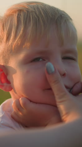un primer plano que captura el tierno momento en que la mano de una madre limpia la cara de su hijo pequeño, centrándose en sus ojos, nariz y boca. el niño, con una expresión seria, está en un campo de hierba durante la puesta del sol