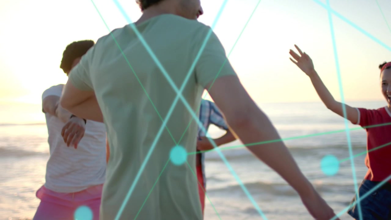 Mann in grünem T-Shirt kommt herein, Freunde begrüßen ihn am Strand bei Sonnenuntergang, türkisfarbene Knoten verbinden ihn mit Technologie.