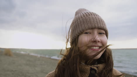 vista de cerca de una adolescente mirando la cámara y sonriendo a la orilla del mar en un día ventoso