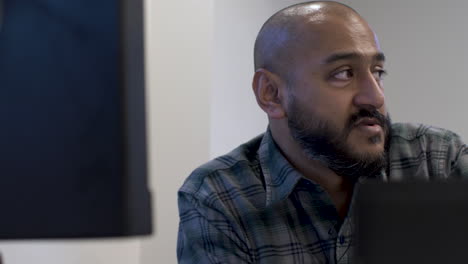 Ethnic-Minority-Bald-Male-Wearing-Green-Cheque-Shirt-Talking-To-Colleague-Across-From-Him