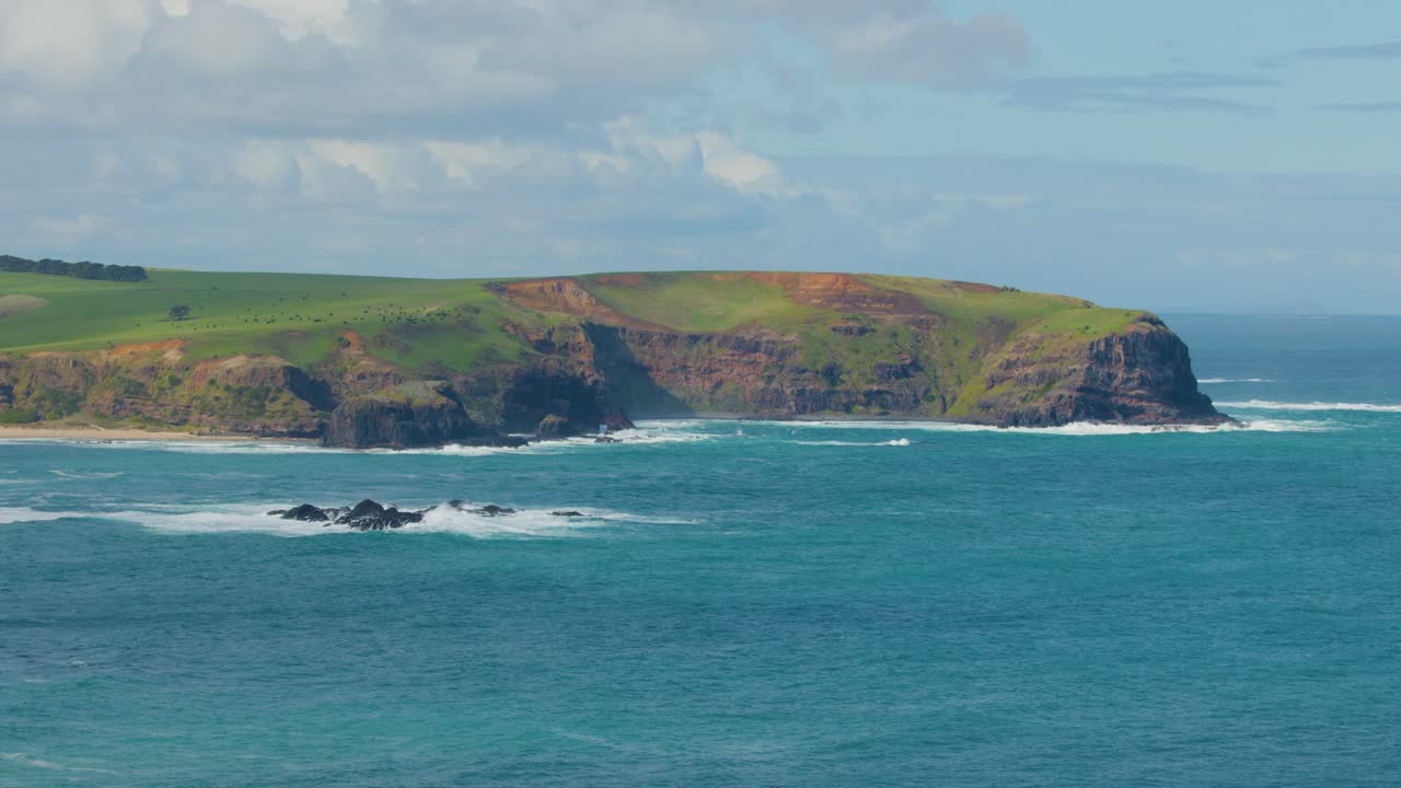 Sweeping view of cliffs, ocean waves, rocky shoreline, and green pastureland under cloudy skies