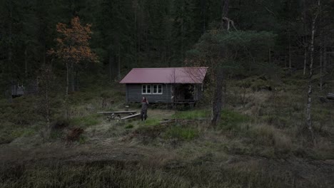 Hildremsvatnet,-Trondelag-County,-Norway---A-Gentleman-Strolling-Toward-the-Lakeshore,-With-a-Fisherman's-Cottage-in-the-Backdrop---Aerial-Pullback