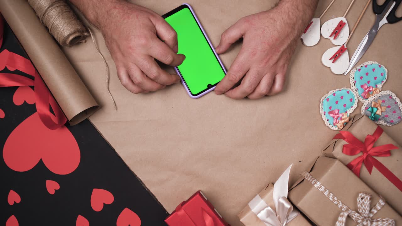 las manos masculinas usan un teléfono inteligente digital con pantalla verde. fondo de mesa con corazones y cajas. concepto de regalo para el día de san valentín