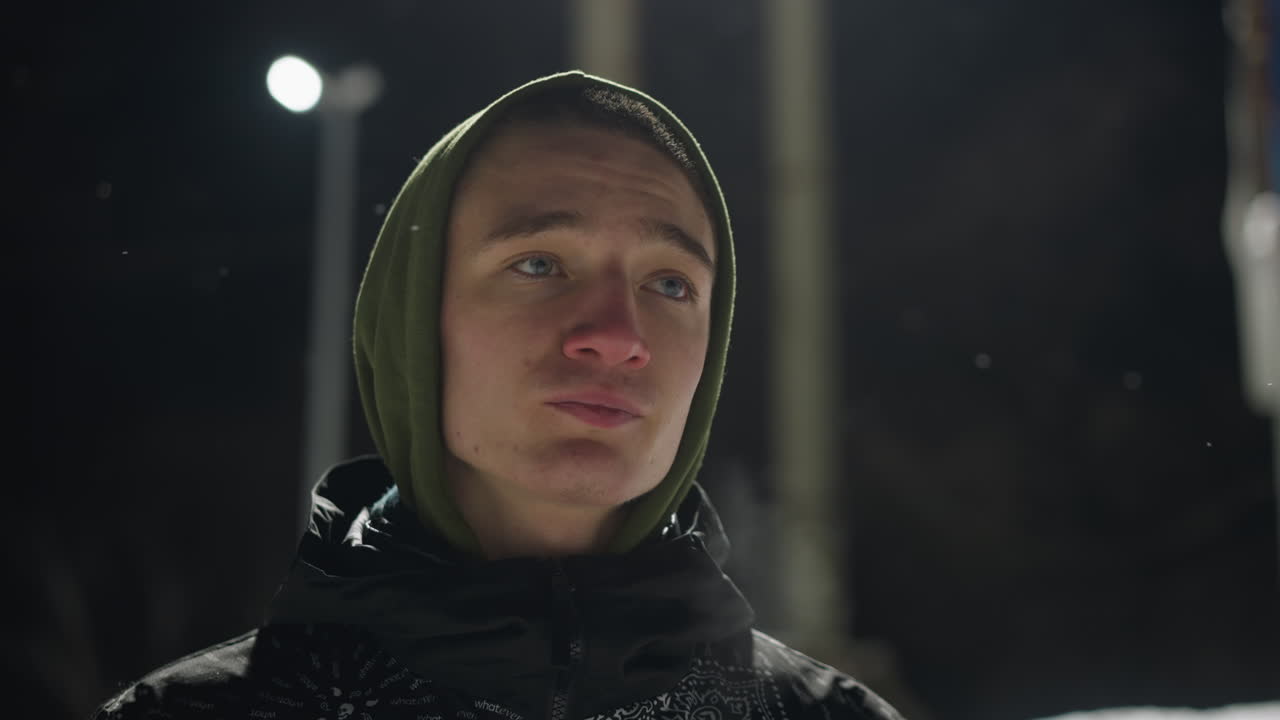 Portrait of teenager in hoodie standing outdoors under intense light during night, with visible breath and snow particles in air, gazing around with quiet, contemplative expression in cold atmosphere