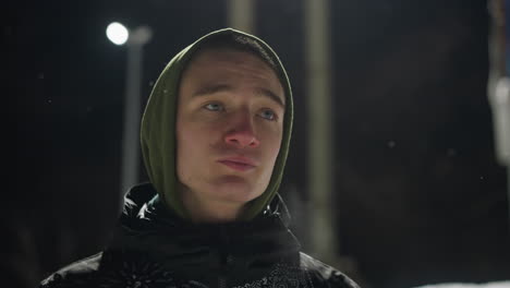portrait of teenager in hoodie standing outdoors under intense light during night, with visible breath and snow particles in air, gazing around with quiet, contemplative expression in cold atmosphere