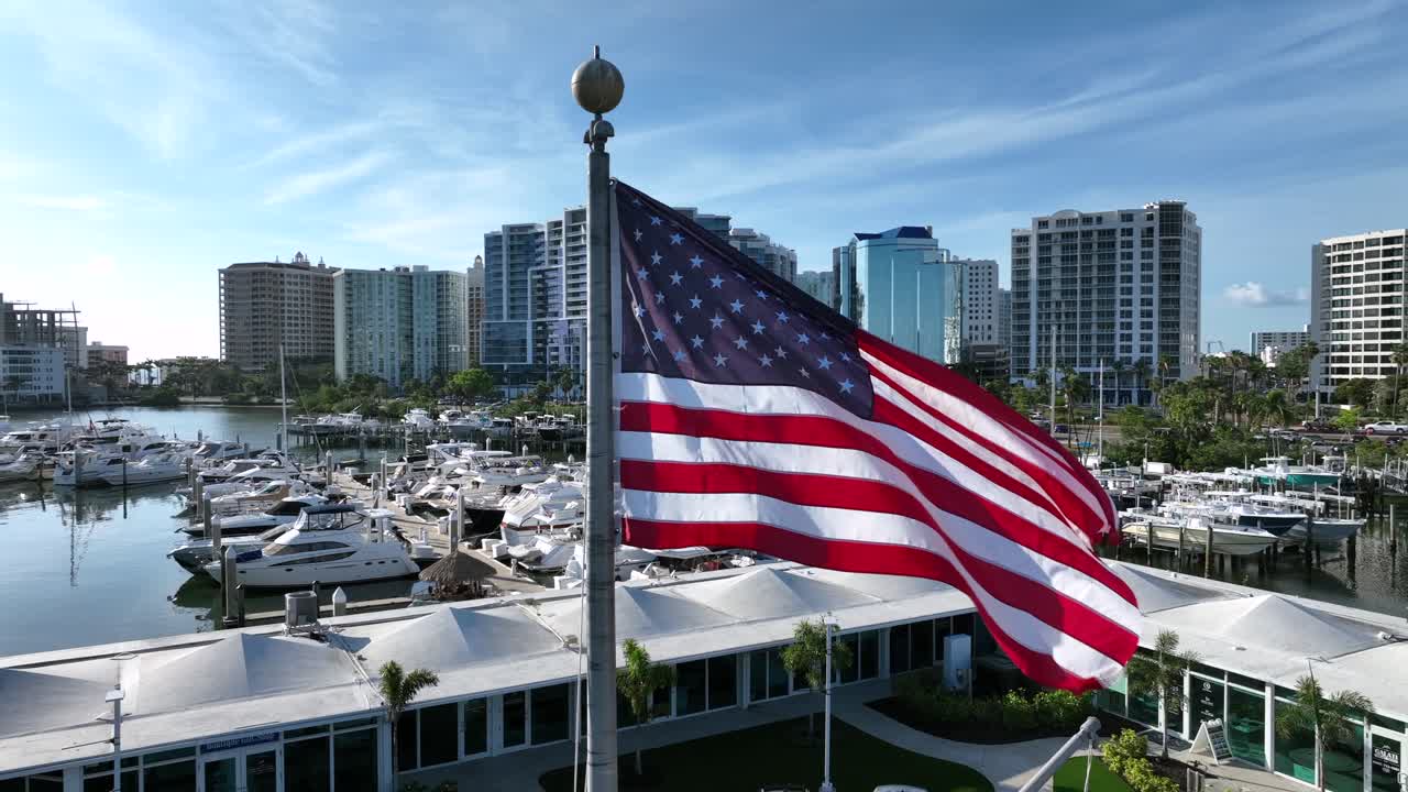 Bandiera degli Stati Uniti d'America che sventola sull'asta della bandiera davanti al porto turistico di Sarasota. Ripresa aerea ravvicinata in orbita. Edifici residenziali e torri del centro sullo sfondo.