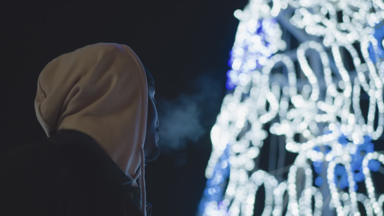 Close-up of man with hoodie looking at glowing blue and white lights in urban city night, holiday season lights. Reflective expression, warm city lights in background, peaceful atmosphere