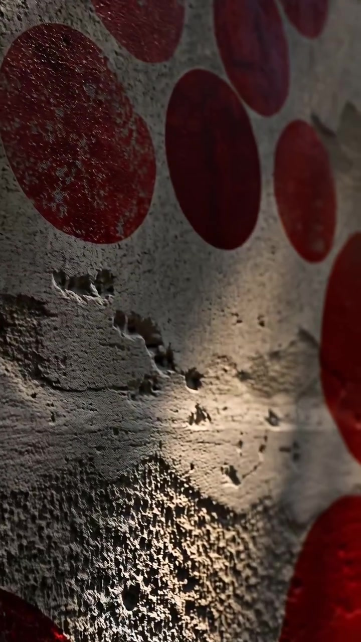 Close perspective moves steadily across textured gray wall covered with distressed red circular marks, while angled light emphasizes peeling paint, holes, scratches, and uneven surface relief.
