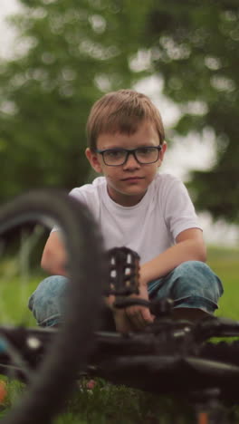 un niño se agacha al lado de su bicicleta en un campo cubierto de hierba, mirando enfocado, con la vista parcial de alguien más de pie cerca