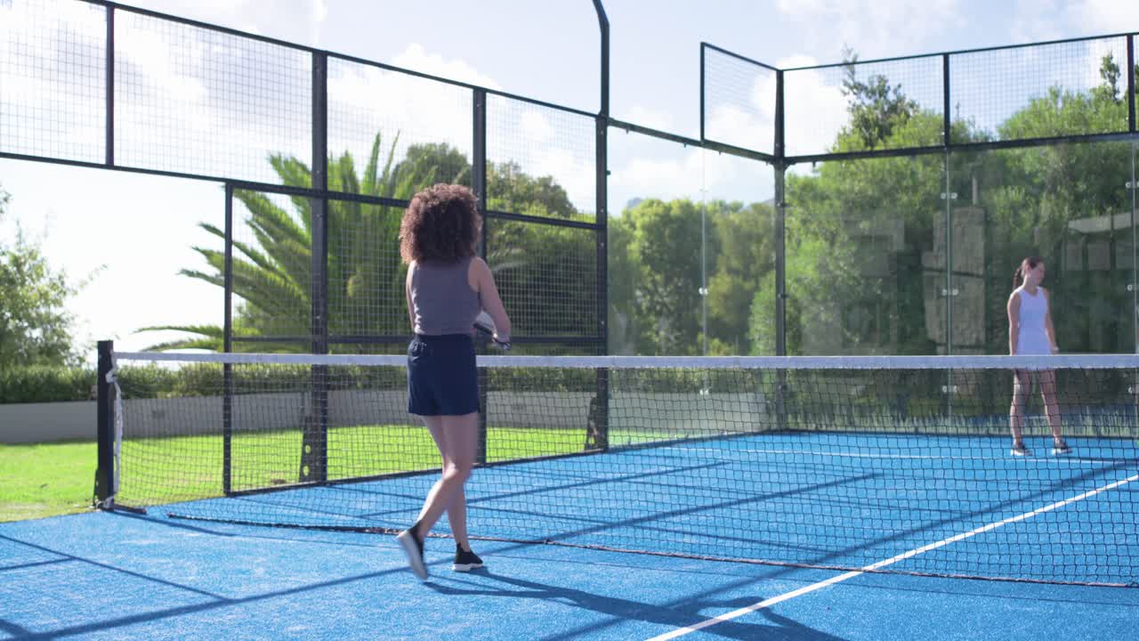 Verschiedene Freundinnen spielen auf einem Padel-Platz; eine Frau mit lockigem Haar beginnt nach einem Punkt einen High-Five mit dem Schläger.