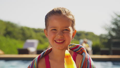 Familia-En-Vacaciones-De-Verano-Con-Una-Chica-Comiendo-Polo-De-Hielo-Al-Borde-De-La-Piscina-Mirando-A-La-Cámara