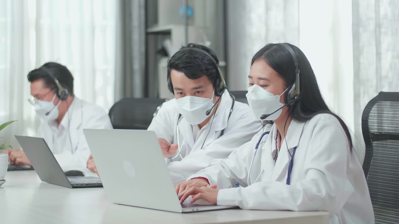 A Man And A Woman Of Three Asian Doctors With Stethoscopes In Headsets And Masks Working As Call Center Agent Discussing Work While Their Colleague Is Speaking On The Call At The Office