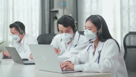 a man and a woman of three asian doctors with stethoscopes in headsets and masks working as call center agent discussing work while their colleague is speaking on the call at the office