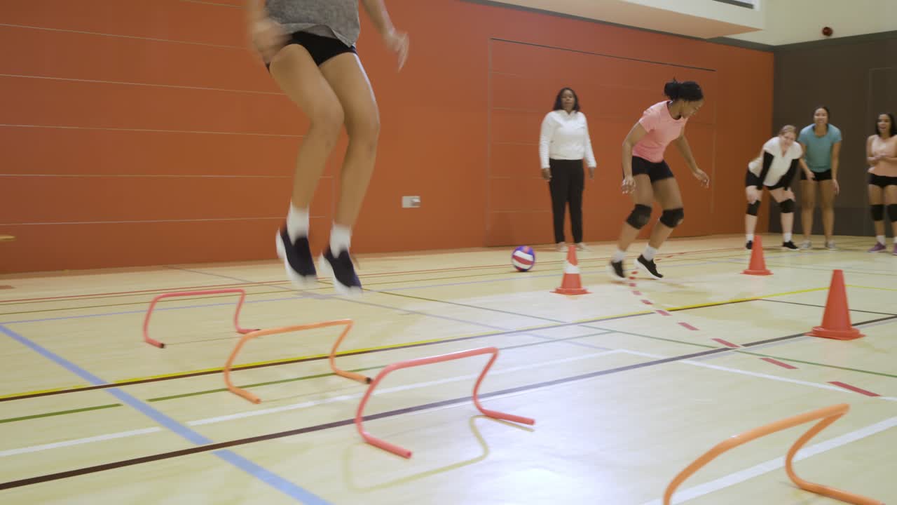 Equipe de vôlei feminino em ginásio observando o treinador; mulher asiática realizando exercícios de agilidade com cones e saltos de barreira para velocidade
