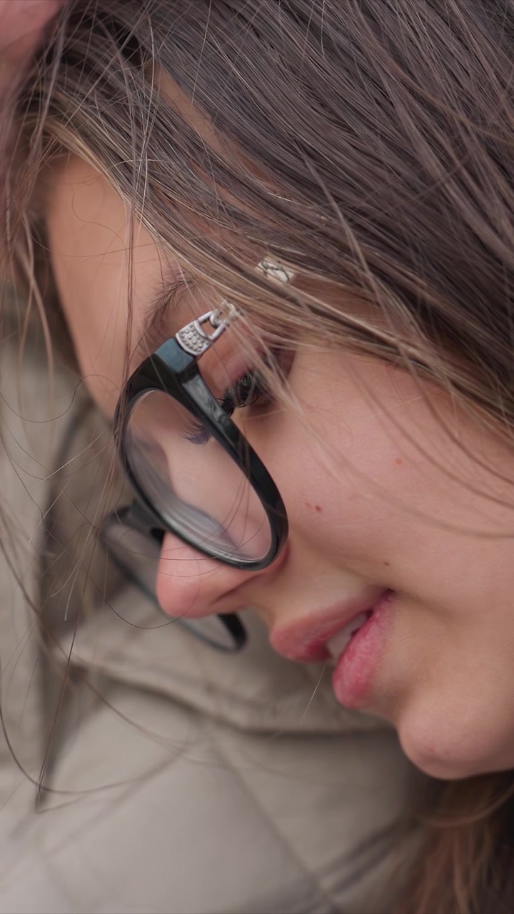 Primo piano di una giovane donna con capelli castani e occhiali neri. La testa è leggermente reclinata verso il basso contro uno sfondo indistinto. L'inquadratura intima enfatizza dettagli come le aste argentate degli occhiali