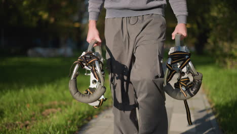 person in dark gray sweatpants and matching hoodie holding metallic stilts with orange and black straps in blurred grassy outdoor area, focused on lower body, showcasing balance and movement