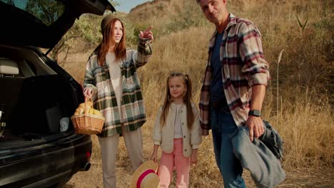 a man with gray hair in a plaid shirt together with his brunette girlfriend in a green plaid shirt with a basket of food and their little daughter are preparing to go on a picnic in a steppe area with dry grass in summer