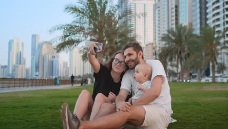 Happy-family-with-two-children-sitting-together-on-grass-in-park-and-taking-a-selfie.-With-smartphone.