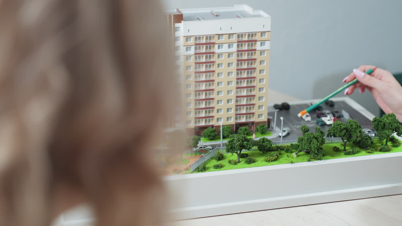 Close up of realtor hand holding pencil and pointing toward detailed building model with visible trees, parked cars, and playground while client attentively observes during office consultation