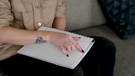 The-Camera-Focuses-On-Woman-Hand-Reading-A-Braille-Book-Sitting-On-The-Sofa-At-Home-2