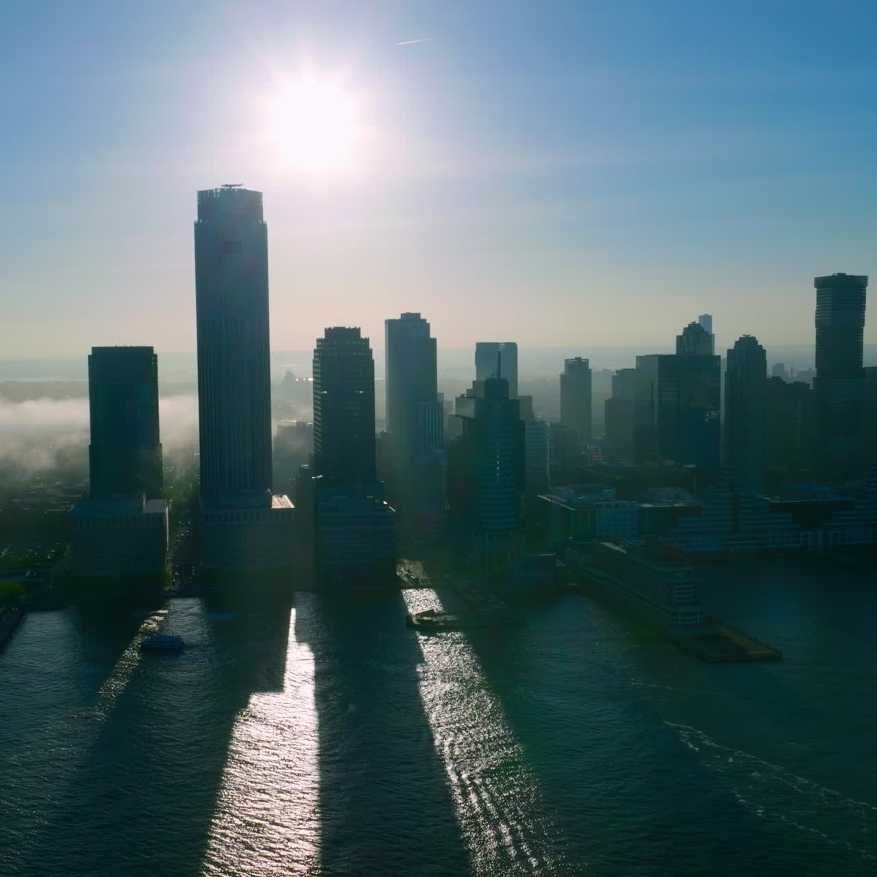 Silhuetas cinzas dos arranha-céus de Nova York contra o sol. Raios de sol passando entre os prédios. Névoa se espalhando pela cidade