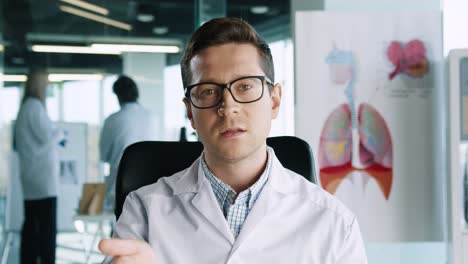 vista de primer plano de un médico caucásico con anteojos sentado en la clínica en el lugar de trabajo mirando la cámara y hablando en consulta médica