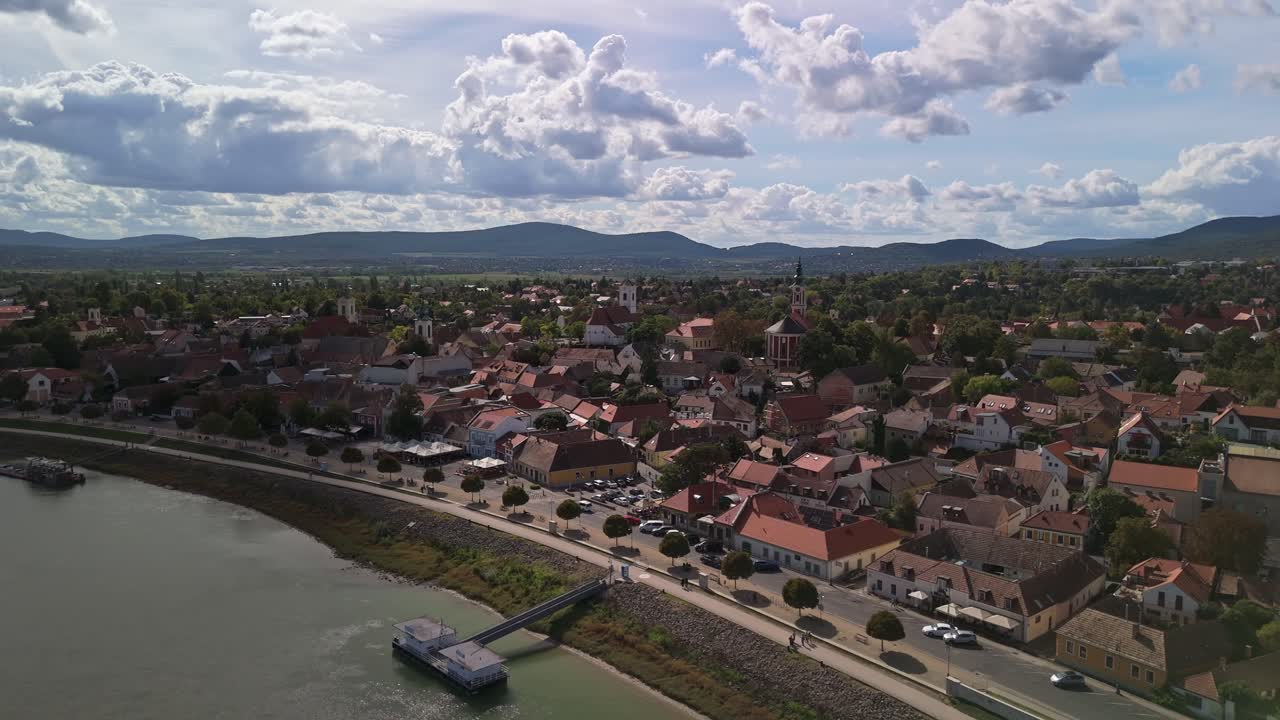 Drone flying forward towards Szentendre, Hungary, revealing the charming red rooftops, Danube River promenade, and surrounding hills under a partly cloudy late afternoon sky