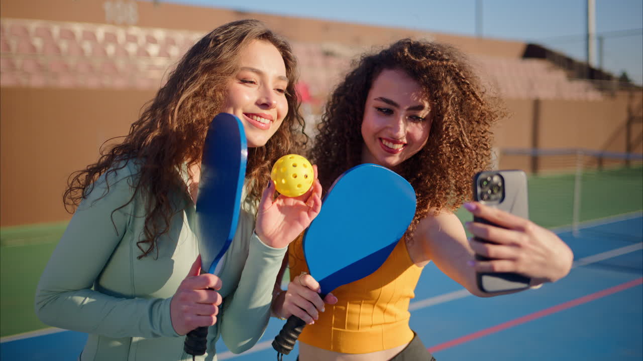 Two girls with curly hair, holding pickleball rackets smiling and talking while taking selfies on a blue court