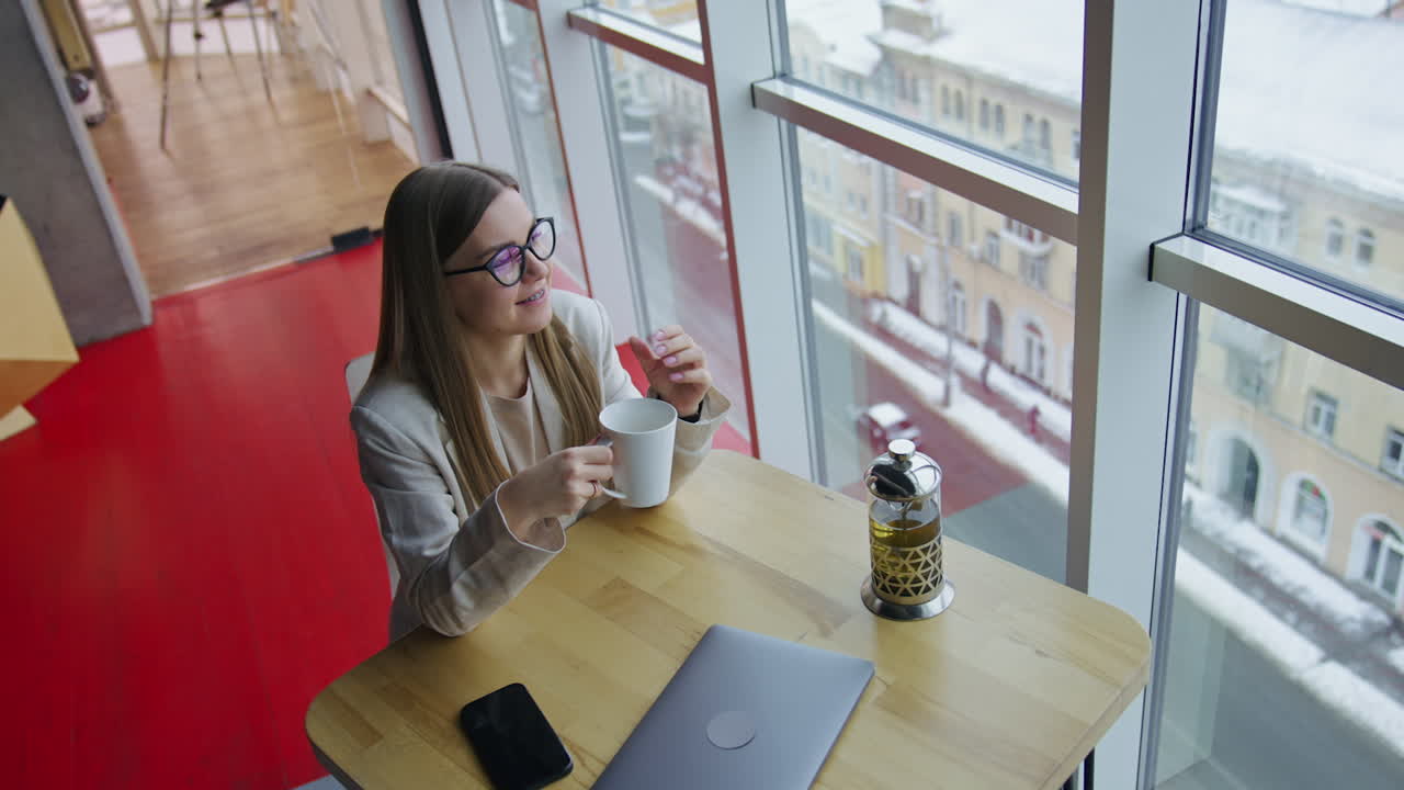 Long-haired young woman having tea sitting at the table. Lady enjoying lunch break and looking out of the window. High angle view.