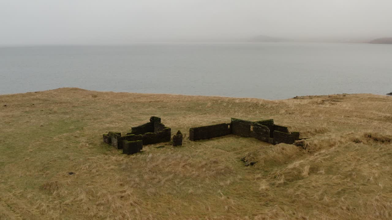 Forladte stenruiner pgra grsklningskystbakke i Hvalfjrur Island med udsigt over roligt hav med knuste murrester og trrt, gyldent terrn i overskyet dagslys