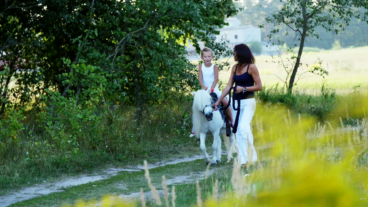 A woman and a boy are walking around the field, son is riding a pony, mother is holding a pony for a bridle. Cheerful, happy family vacation. Outdoors, in summer, near the forest