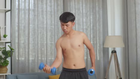 아시아 십대 소년 은 집 에서 셔츠 를 벗고 운동 하는 동안 덤벨 으로 훈련 하고 있다