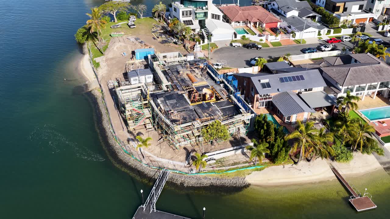 Drone footage captures a sunny, high-angle view of residential house construction along a canal in Gold Coast, Australia, with smooth lateral camera movement
