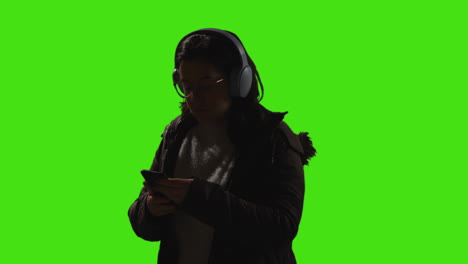 woman wearing wireless headphones streaming music from mobile phone against green screen with low key lighting 2