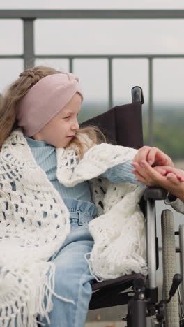 loving mom gently strokes small hand of daughter with disability. woman and little girl covered with shawl in wheelchair on observation deck overlooking river