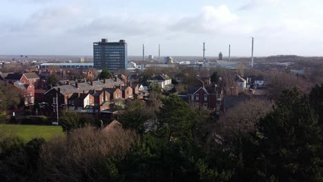 Aerial-view-over-park-trees-with-birds-flying-through-scene-to-industrial-townscape-with-blue-skyscraper,-Merseyside,-England