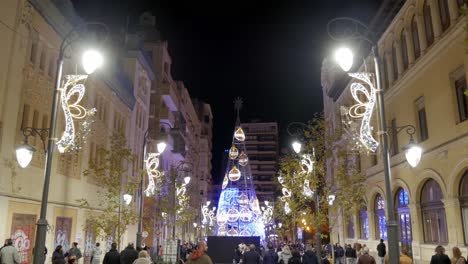 play of lights on a giant christmas tree in the center of the street surrounded by buildings with people walking around in alicante, spain at night. avenia de la constitucion.