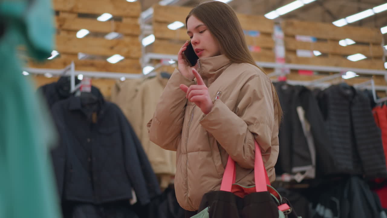 A young girl wearing a light brown jacket is standing in a clothing mall, holding a phone to her ear while talking and explaining with her hand. She is carrying a gym bag with bright pink straps