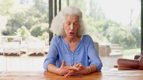 senior woman in blue shirt on video call at table, talking and gesturing with hands